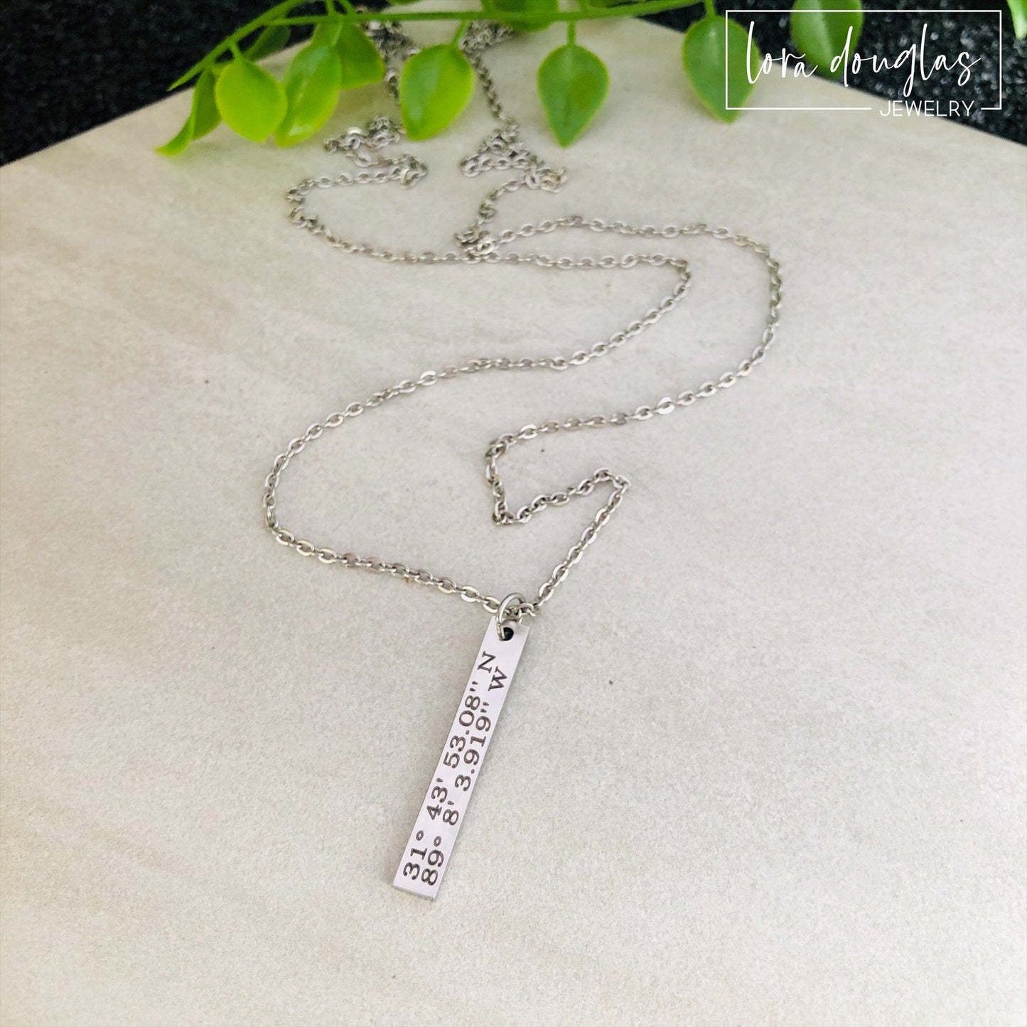 A custom coordinates necklace with a silver stainless steel pendant and chain, displayed against a floral background.