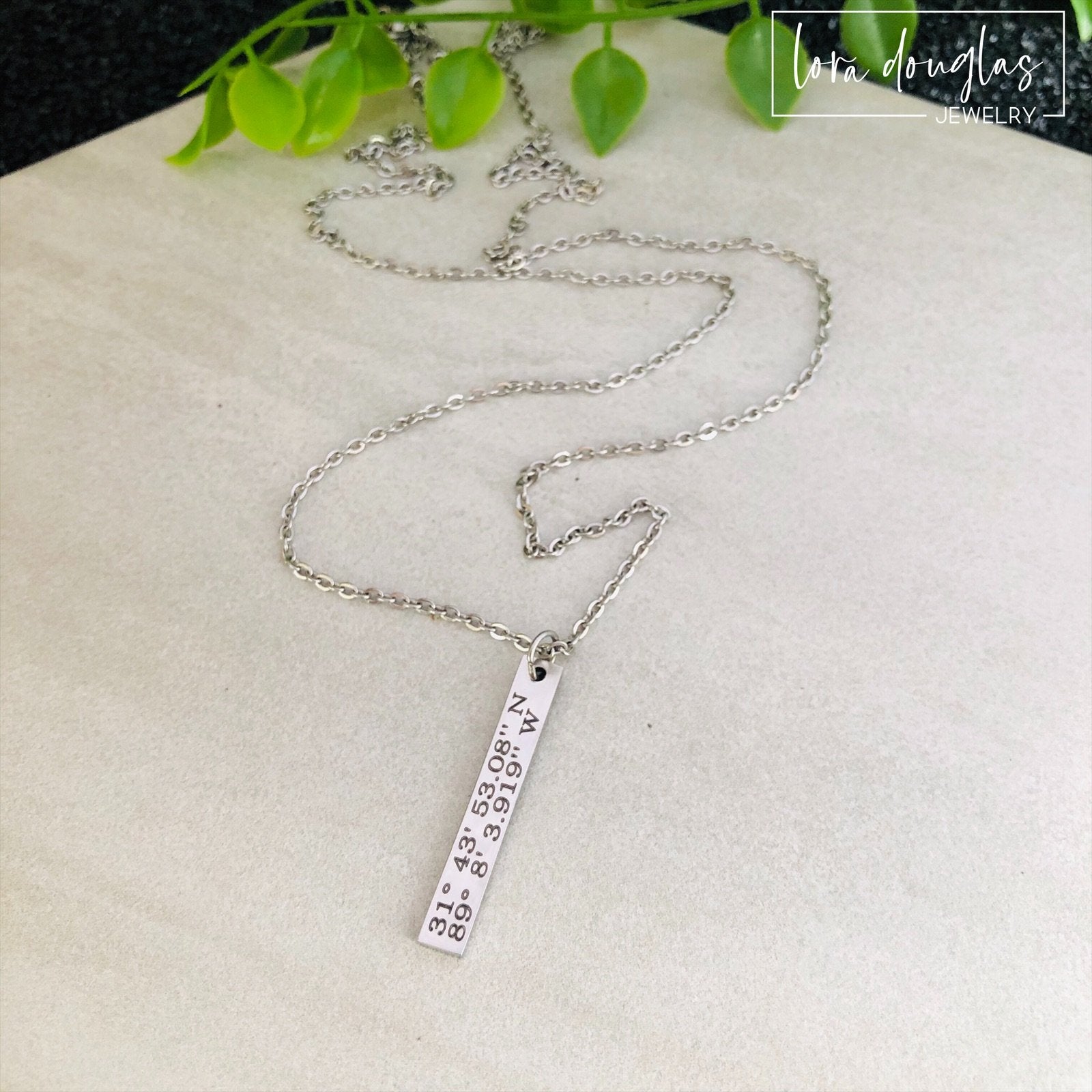 A custom coordinates necklace with a silver stainless steel pendant and chain, displayed against a floral background.