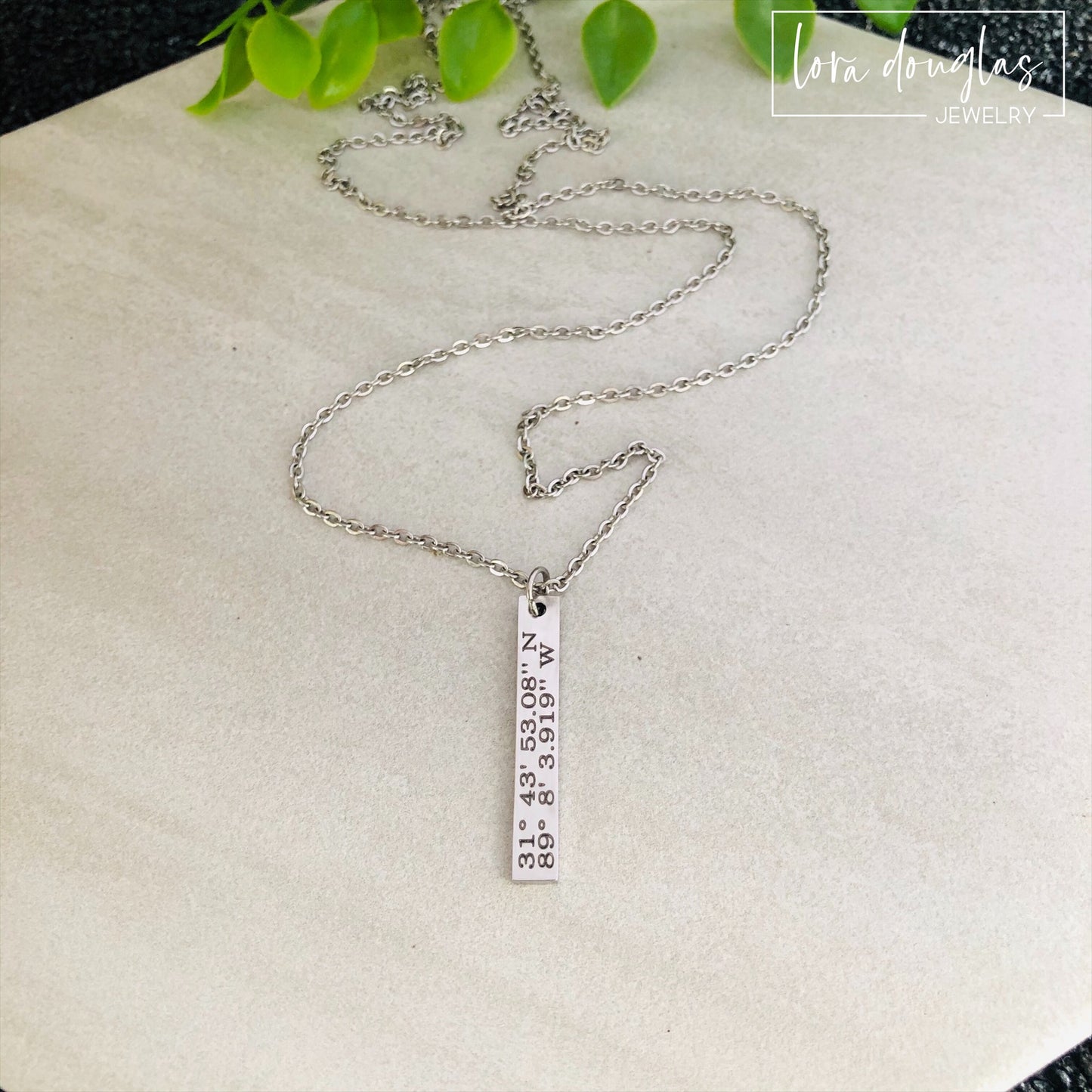 A custom coordinates necklace with a silver stainless steel pendant and chain, displayed against a floral background.