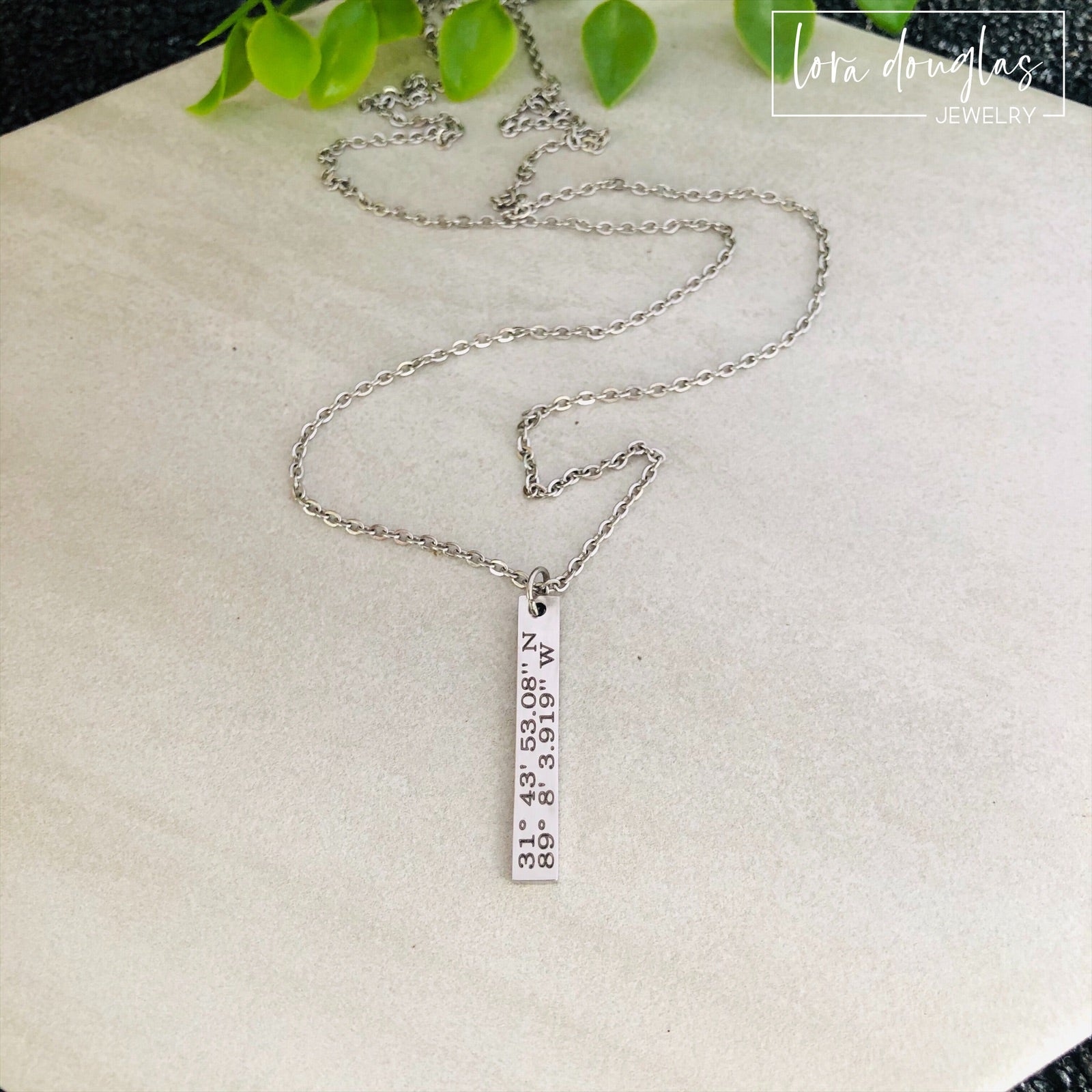 A custom coordinates necklace with a silver stainless steel pendant and chain, displayed against a floral background.