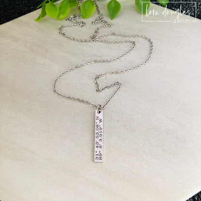 A custom coordinates necklace with a silver stainless steel pendant and chain, displayed against a floral background.
