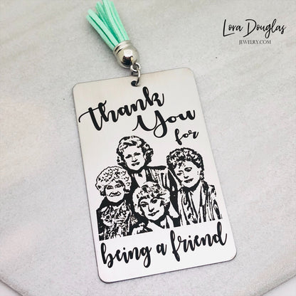 A stainless steel bookmark with engraved text and illustrations of the Golden Girls characters, with a navy blue velvet drawstring pouch and a gift tag.