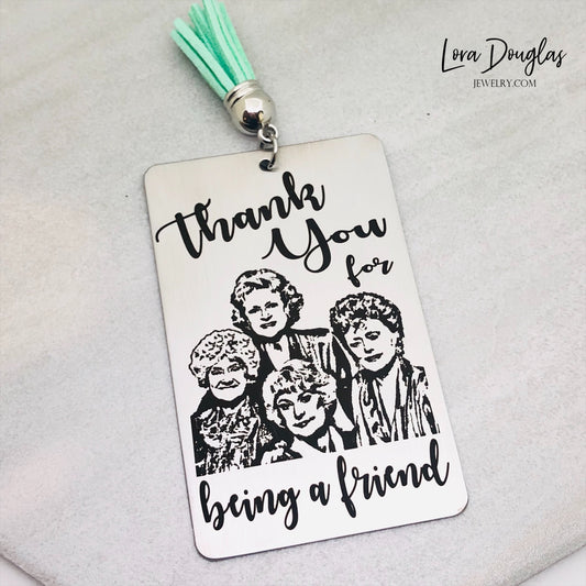 A stainless steel bookmark with engraved text and illustrations of the Golden Girls characters, with a navy blue velvet drawstring pouch and a gift tag.