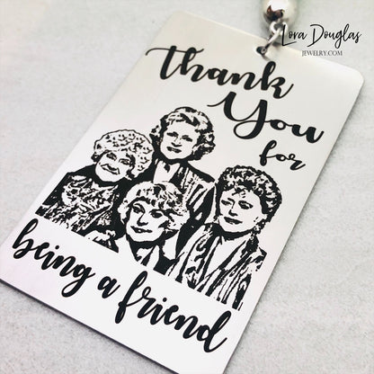 A stainless steel bookmark with engraved text and illustrations of the Golden Girls characters, with a navy blue velvet drawstring pouch and a gift tag.