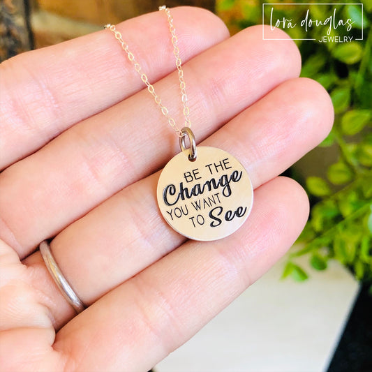 A round silver pendant with engraved text 'BE THE CHANGE YOU WANT TO SEE' displayed on a chain.