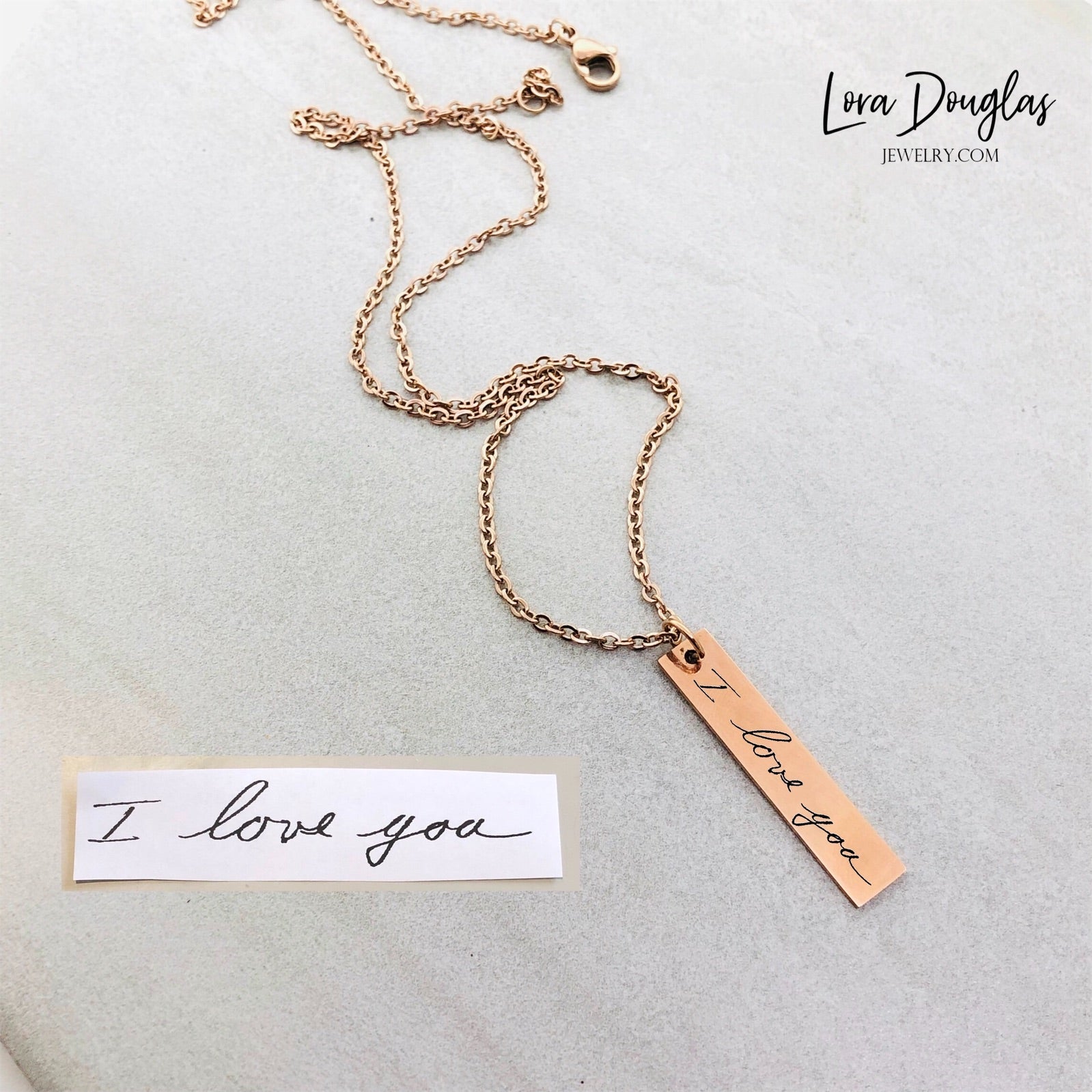 A gold stainless steel pendant with engraved handwriting, attached to an 18 inch chain with a lobster clasp, displayed against a white background with a blurred background hinting at a floral arrangement.