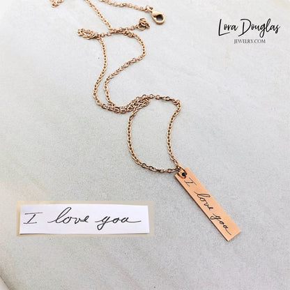 A gold stainless steel pendant with engraved handwriting, attached to an 18 inch chain with a lobster clasp, displayed against a white background with a blurred background hinting at a floral arrangement.