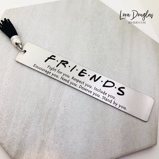 A stainless steel bookmark with an engraved message that reads 'Friends' and includes a tassel, displayed on a textured background.