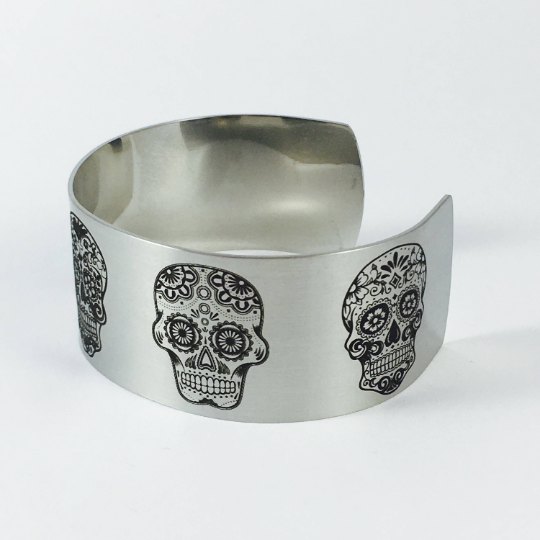 Silver and gold sugar skull cuff bracelets showing the available stainless steel finish options, each featuring five permanently engraved skull designs across a wide 1 inch band with a distinctive matte surface.
