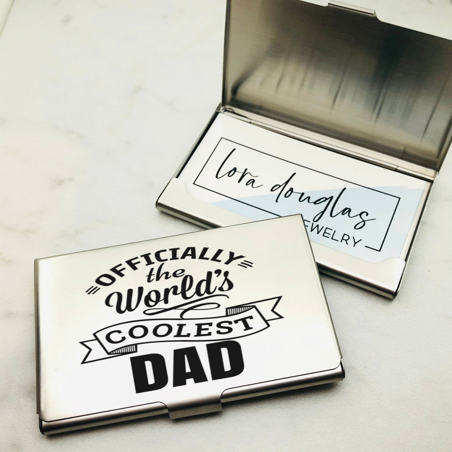 A stainless steel business card holder with the engraving 'OFFICIALLY THE WORLD'S COOLEST DAD' on the front cover, alongside a closed case.
