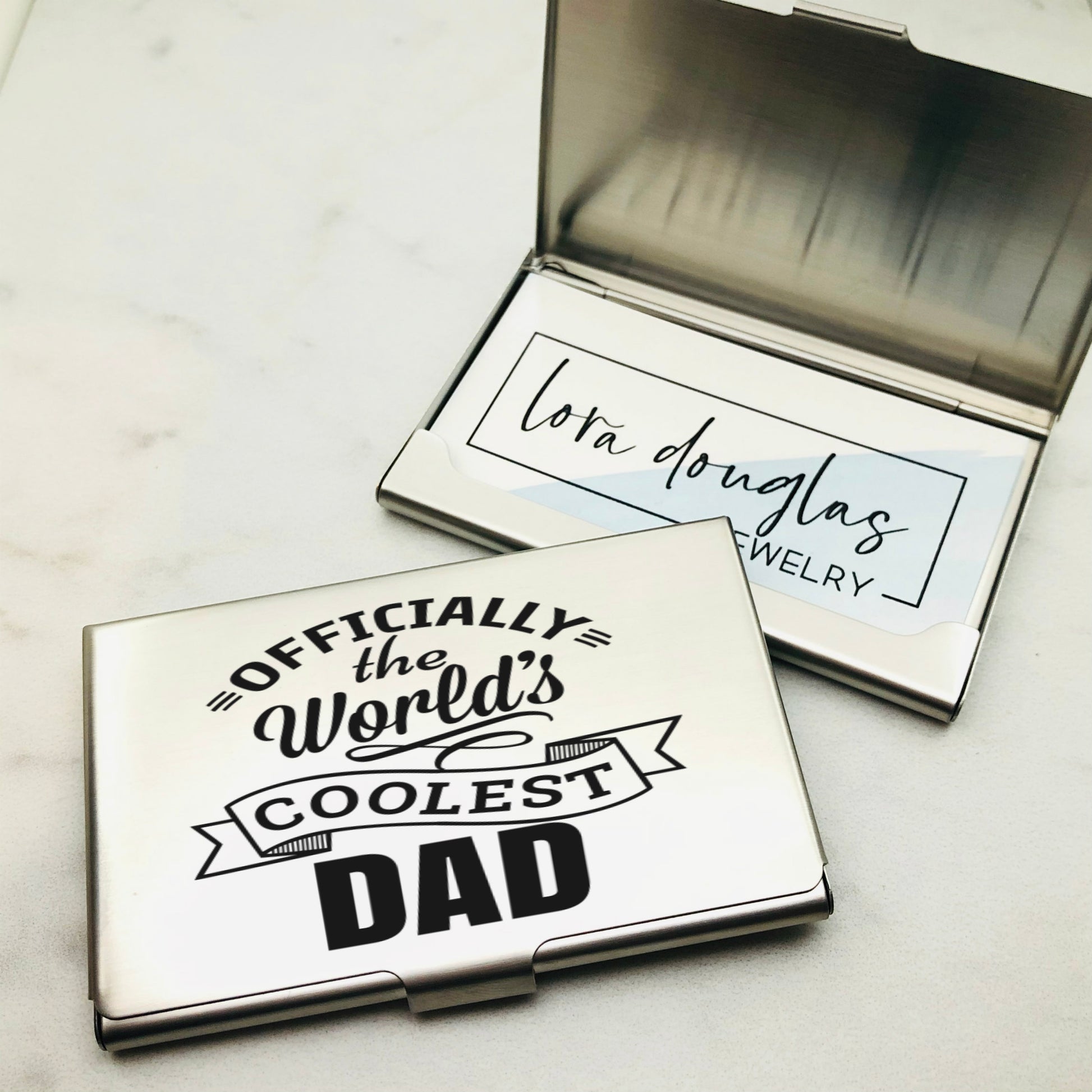 A stainless steel business card holder with the engraving 'OFFICIALLY THE WORLD'S COOLEST DAD' on the front cover, alongside a closed case.