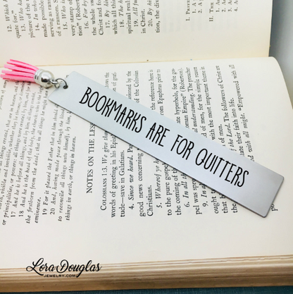 A stainless steel bookmark with the inscription 'BOOKMARKS ARE FOR QUITTERS' engraved on it, displayed on an open book. The bookmark has a tassel on one end.