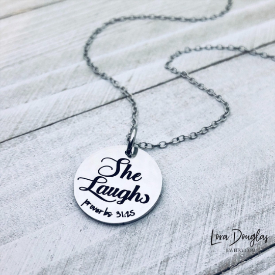 A silver pendant with the inscription 'She Laughs, Proverbs 31:25' on a chain, placed on a wooden surface.