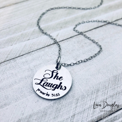 A silver pendant with the inscription 'She Laughs, Proverbs 31:25' on a chain, placed on a wooden surface.