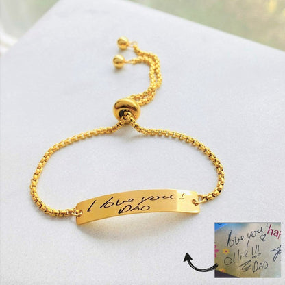 A gold bracelet with a bar pendant engraved with the handwritten message 
