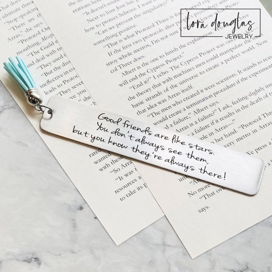 A stainless steel bookmark with an engraved message 'Good friends are like stars, you don't always see them but you know they're there.' placed on a book page, accompanied by a navy blue velvet drawstring pouch with a gift tag.