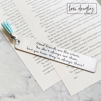 A stainless steel bookmark with an engraved message 'Good friends are like stars, you don't always see them but you know they're there.' placed on a book page, accompanied by a navy blue velvet drawstring pouch with a gift tag.