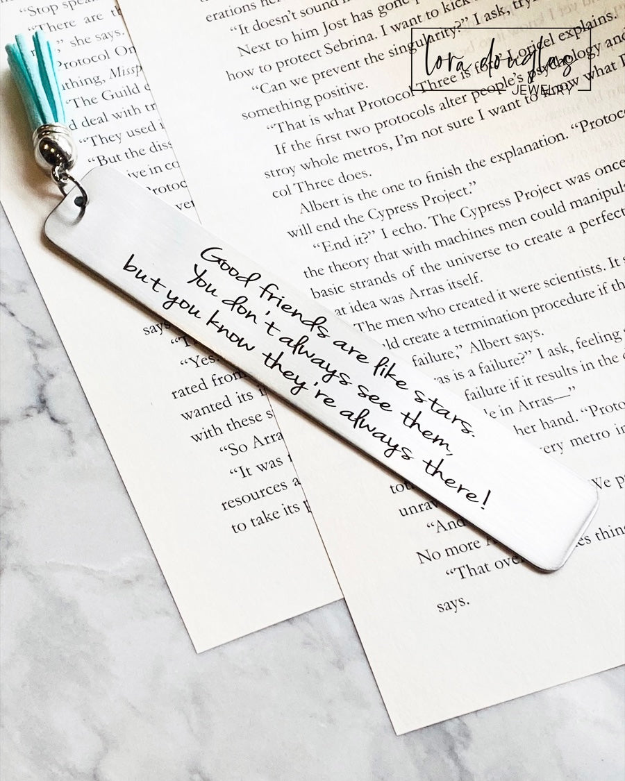 A stainless steel bookmark with an engraved message 'Good friends are like stars, you don't always see them but you know they're there.' placed on a book page, accompanied by a navy blue velvet drawstring pouch with a gift tag.