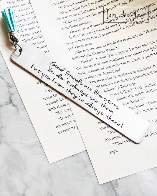 A stainless steel bookmark with an engraved message 'Good friends are like stars, you don't always see them but you know they're there.' placed on a book page, accompanied by a navy blue velvet drawstring pouch with a gift tag.