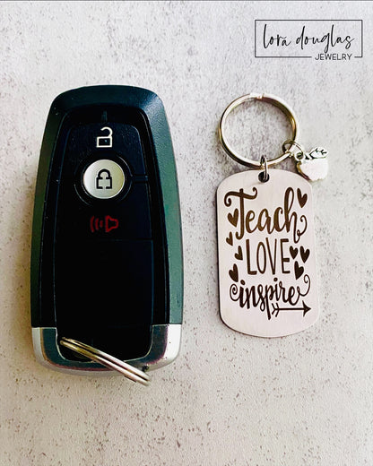 A silver stainless steel dog tag keychain with a silver pewter apple charm, engraved with the text 'Teach Love Inspire' accompanied by a heart symbol.