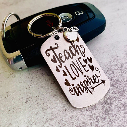 A silver stainless steel dog tag keychain with a silver pewter apple charm, engraved with the text 'Teach Love Inspire' accompanied by a heart symbol.