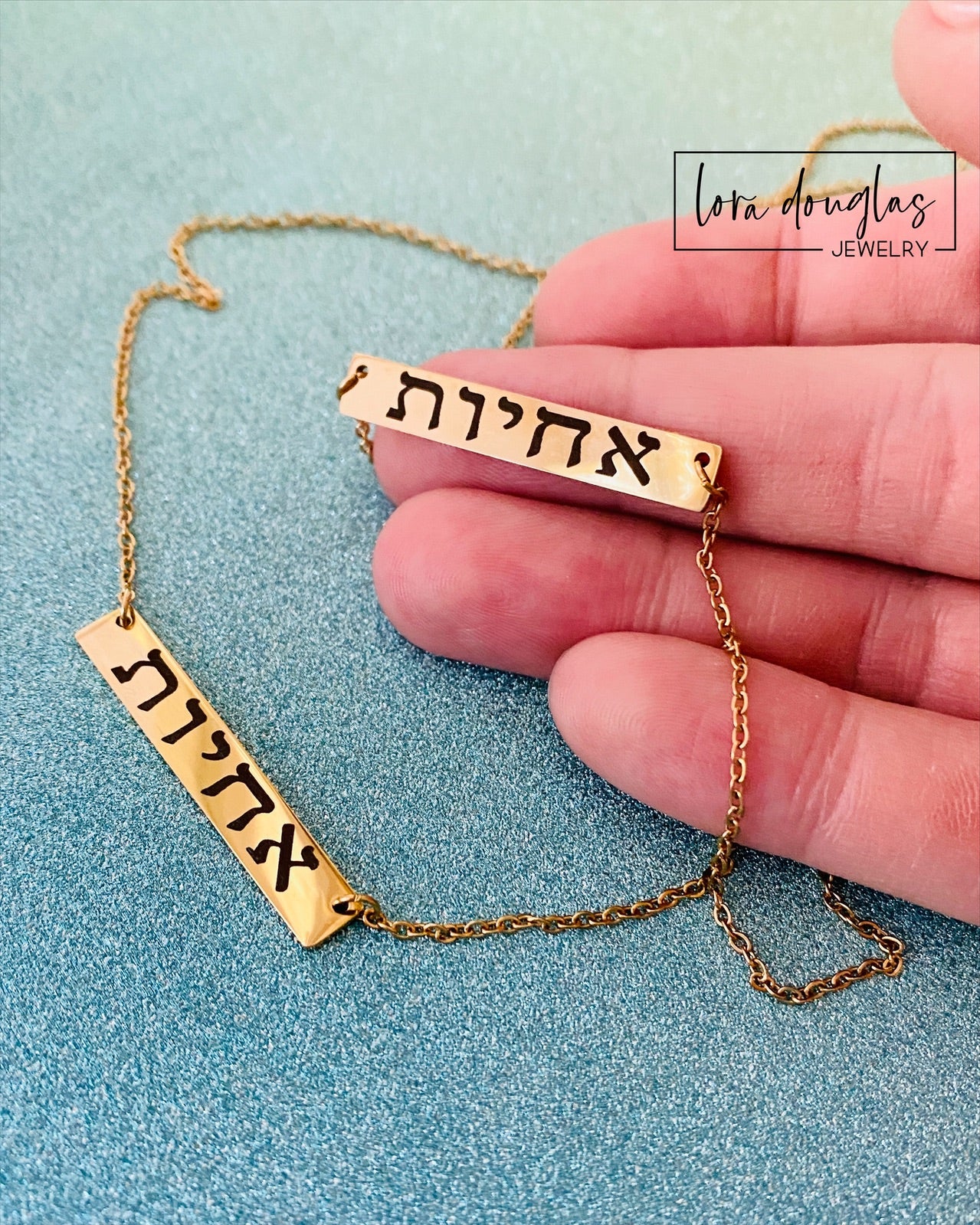 A gold bar necklace engraved with 'SISTERS IN CHRIST' in Hebrew script, presented in a box with the text 'SISTERS IN CHRIST ARE SISTERS FOR LIFE'. 