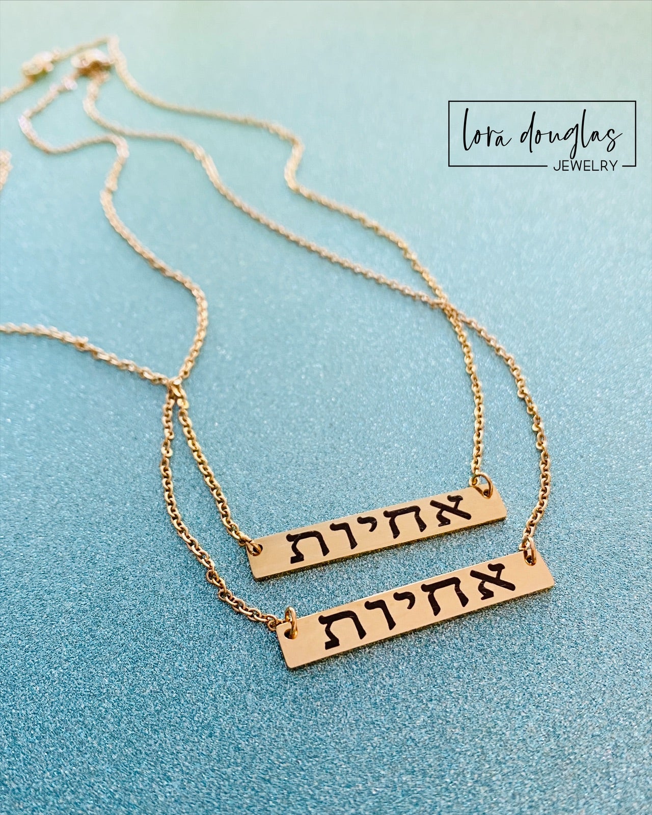 A gold bar necklace engraved with 'SISTERS IN CHRIST' in Hebrew script, presented in a box with the text 'SISTERS IN CHRIST ARE SISTERS FOR LIFE'. 