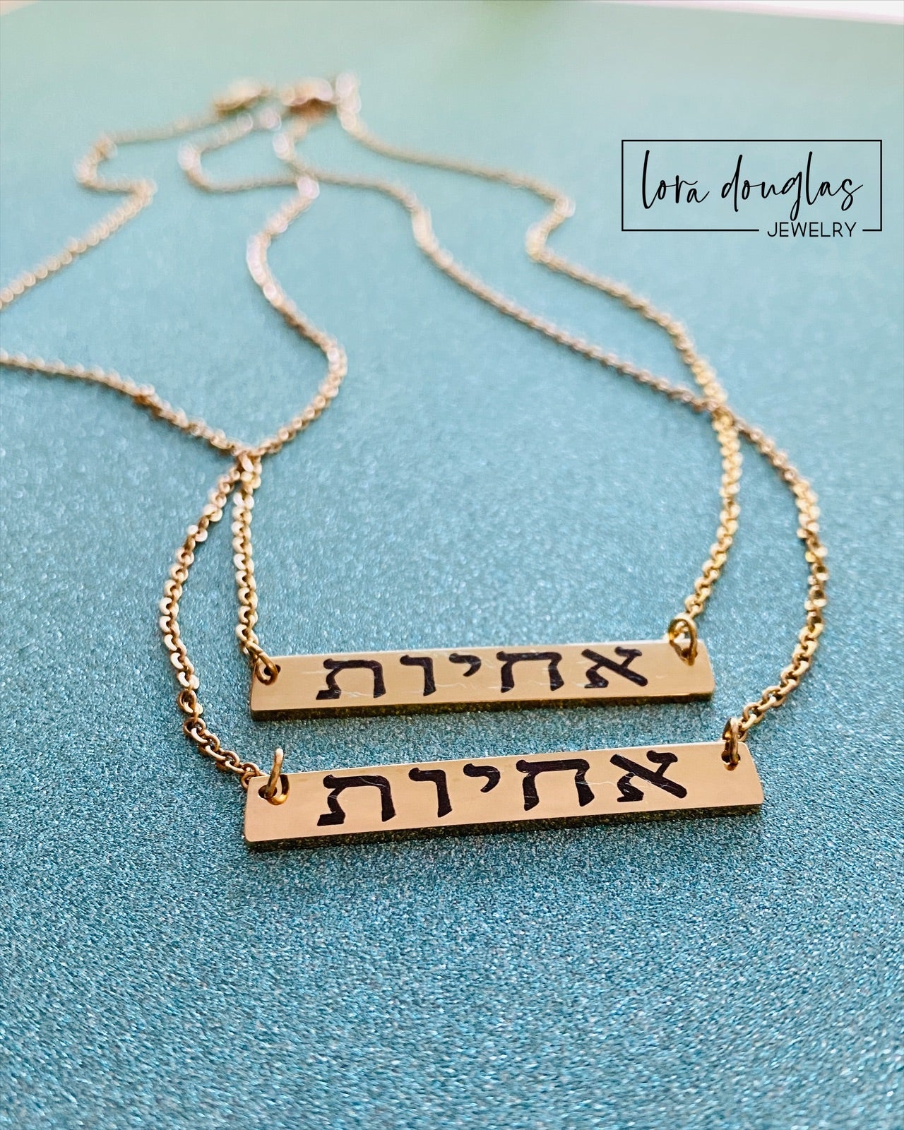 A gold bar necklace engraved with 'SISTERS IN CHRIST' in Hebrew script, presented in a box with the text 'SISTERS IN CHRIST ARE SISTERS FOR LIFE'. 