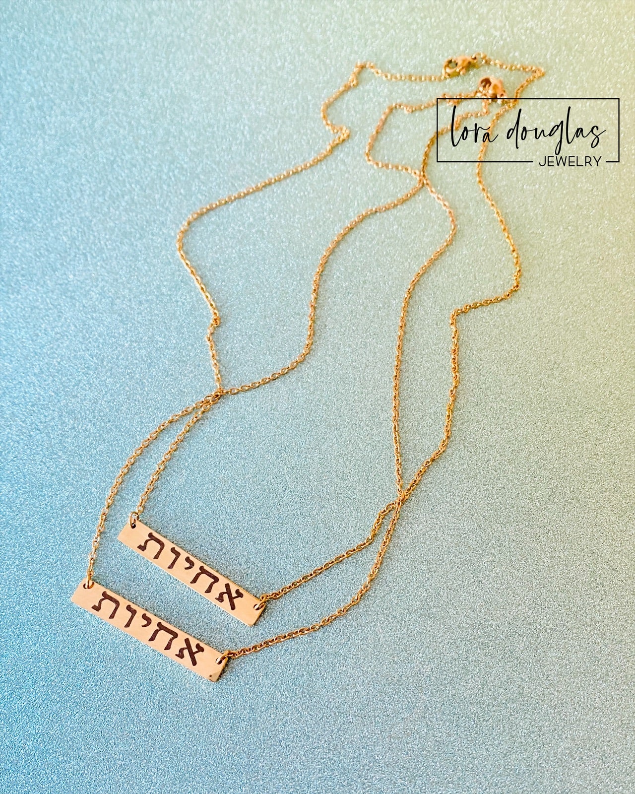 A gold bar necklace engraved with 'SISTERS IN CHRIST' in Hebrew script, presented in a box with the text 'SISTERS IN CHRIST ARE SISTERS FOR LIFE'. 