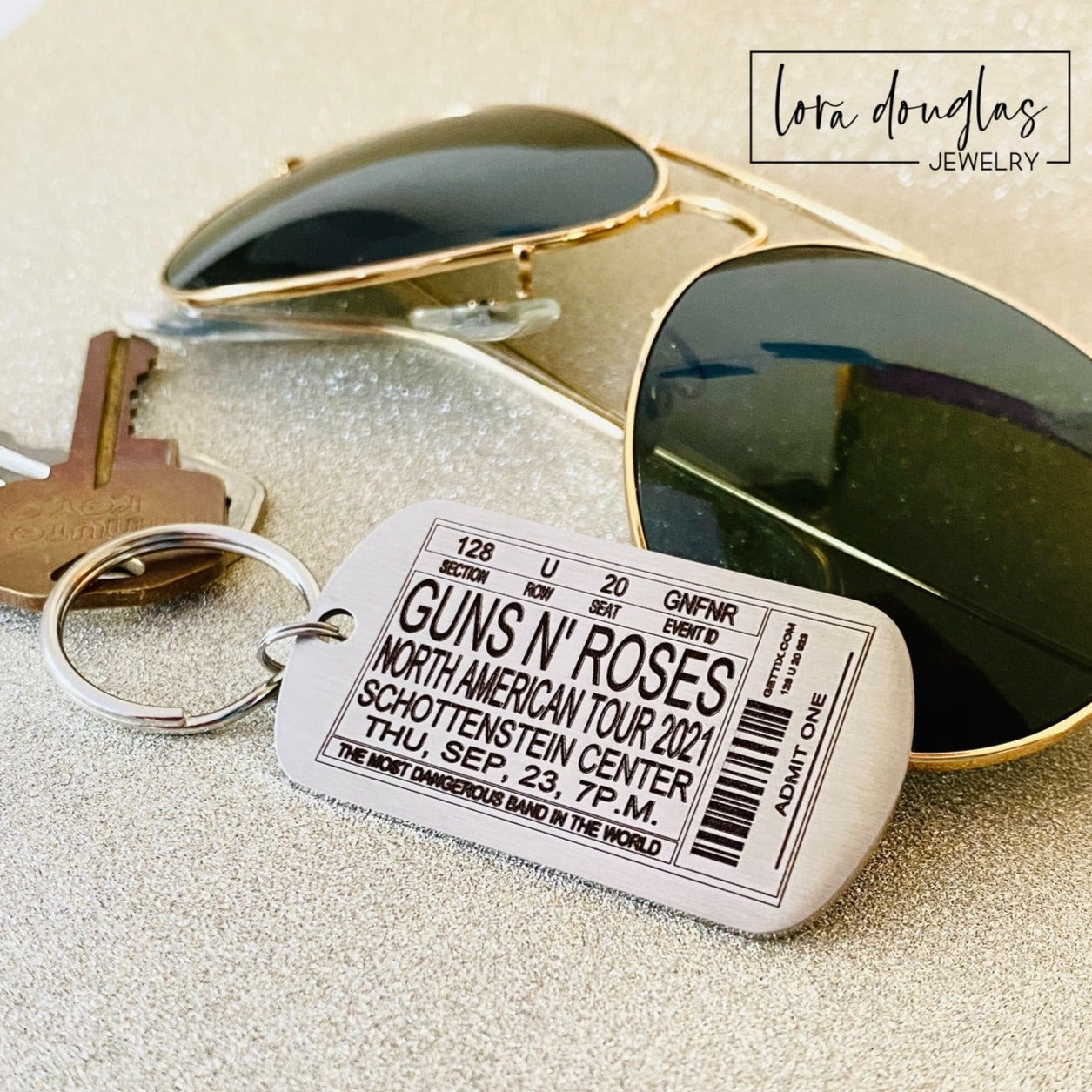 A stainless steel key chain with a concert ticket engraving, accompanied by a pair of sunglasses and a key, on a reflective surface.