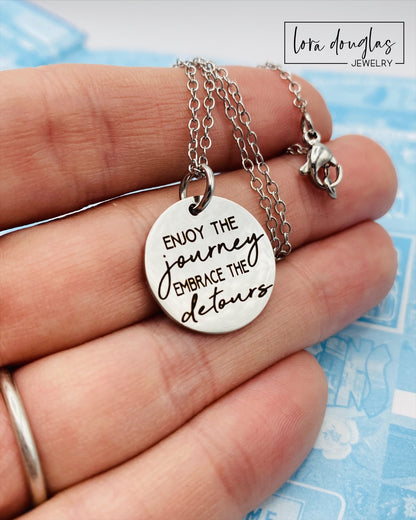 A silver necklace with a lobster clasp and a stainless steel bangle bracelet, both featuring an engraved message 'Enjoy the Journey, Embrace the Detours'