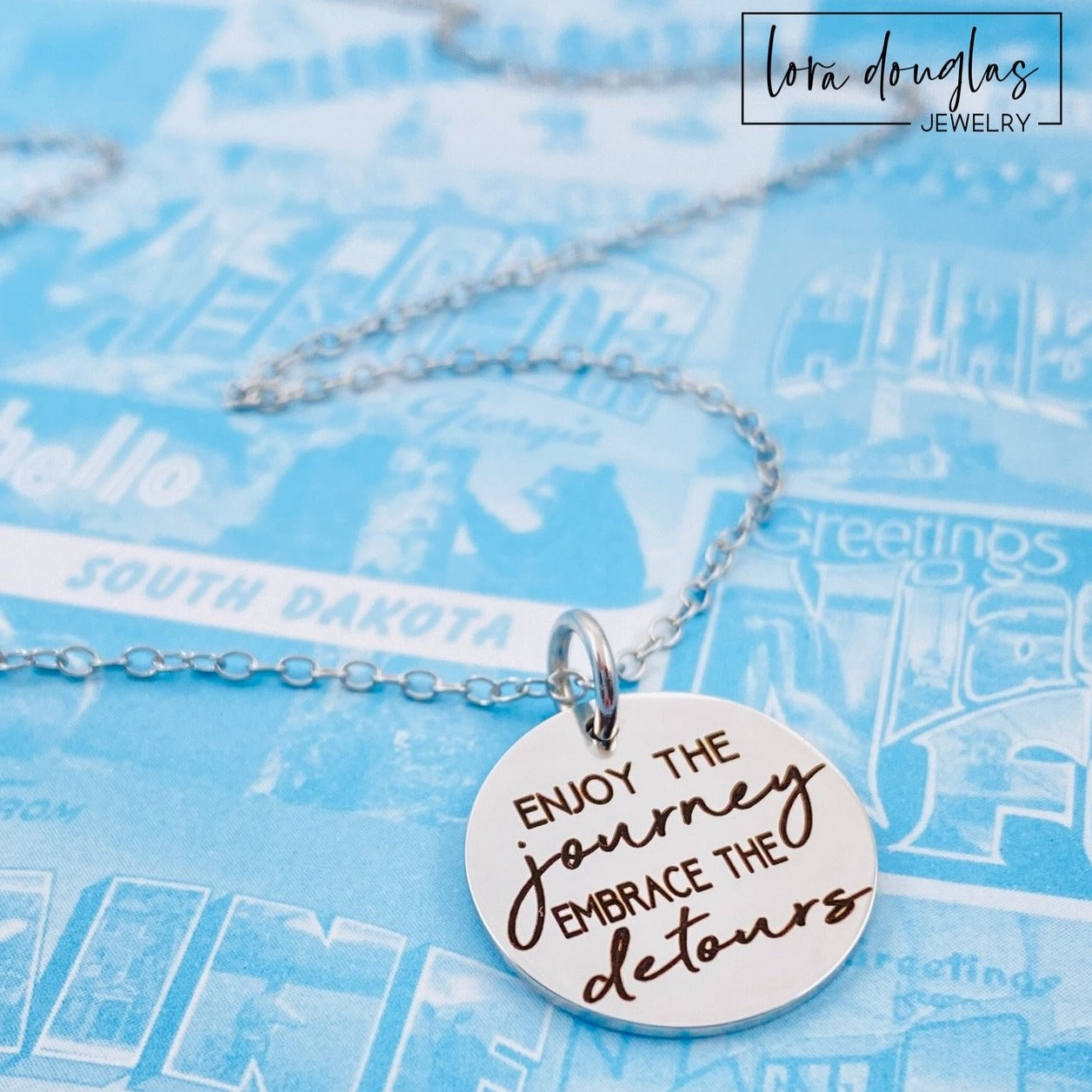 A silver necklace with a lobster clasp and a stainless steel bangle bracelet, both featuring an engraved message 'Enjoy the Journey, Embrace the Detours'