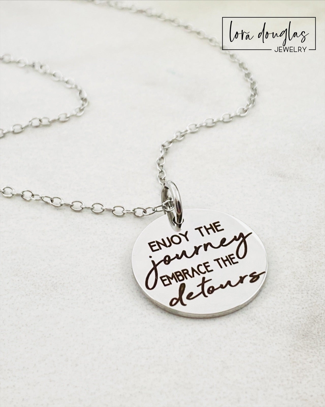 A silver necklace with a lobster clasp and a stainless steel bangle bracelet, both featuring an engraved message 'Enjoy the Journey, Embrace the Detours'