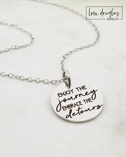 A silver necklace with a lobster clasp and a stainless steel bangle bracelet, both featuring an engraved message 'Enjoy the Journey, Embrace the Detours'