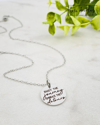 A silver necklace with a lobster clasp and a stainless steel bangle bracelet, both featuring an engraved message 'Enjoy the Journey, Embrace the Detours'