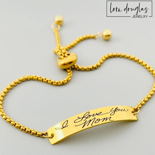 A gold bracelet with a bar pendant engraved with a handwritten message 