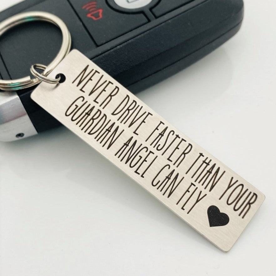Stainless steel keychain with an engraving that reads 'NEVER DRIVE FASTER THAN YOUR Guardian Angel Can Fly' alongside a heart symbol, placed next to a key.