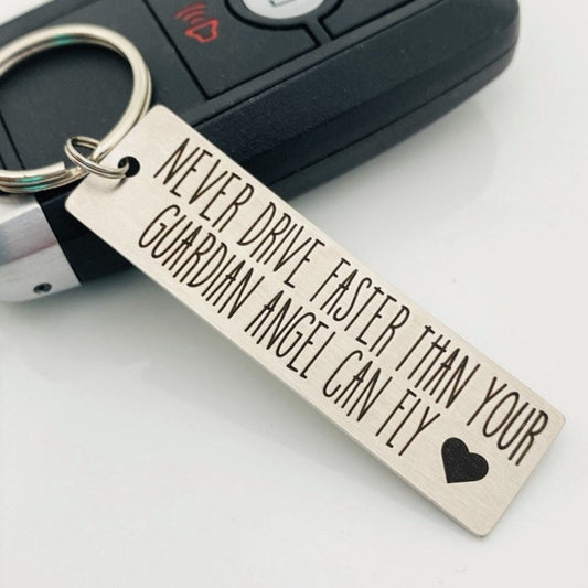 Stainless steel keychain with an engraving that reads 'NEVER DRIVE FASTER THAN YOUR Guardian Angel Can Fly' alongside a heart symbol, placed next to a key.