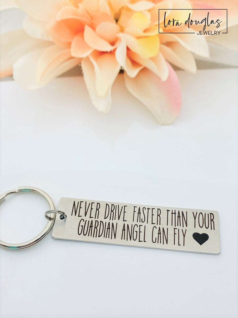Stainless steel keychain with an engraving that reads 'NEVER DRIVE FASTER THAN YOUR Guardian Angel Can Fly' alongside a heart symbol, placed next to a key.