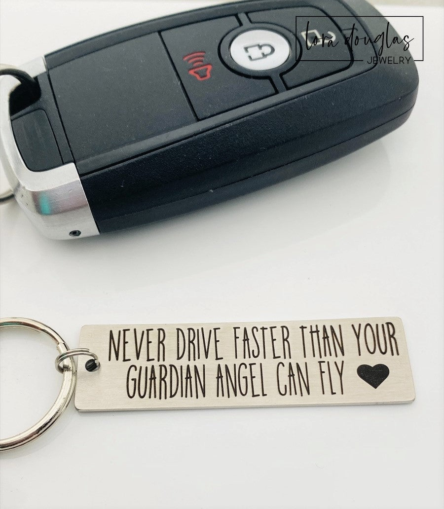 Stainless steel keychain with an engraving that reads 'NEVER DRIVE FASTER THAN YOUR Guardian Angel Can Fly' alongside a heart symbol, placed next to a key.