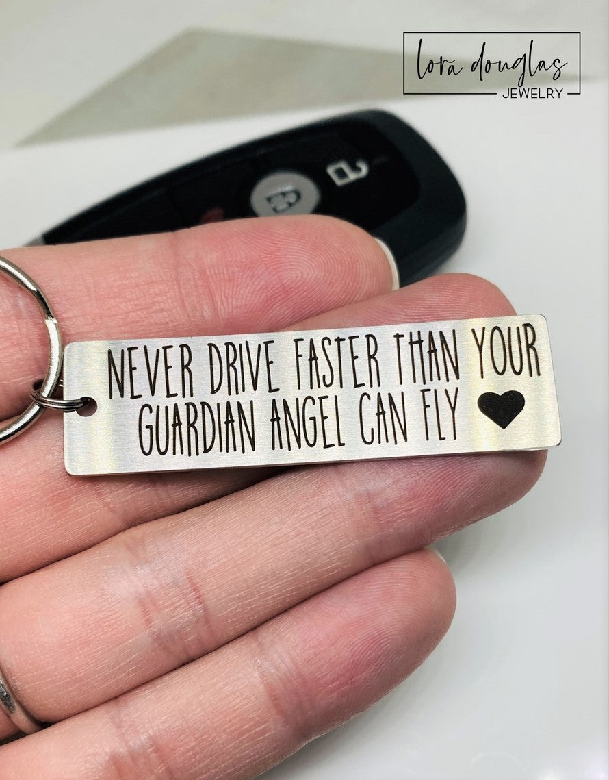 Stainless steel keychain with an engraving that reads 'NEVER DRIVE FASTER THAN YOUR Guardian Angel Can Fly' alongside a heart symbol, placed next to a key.