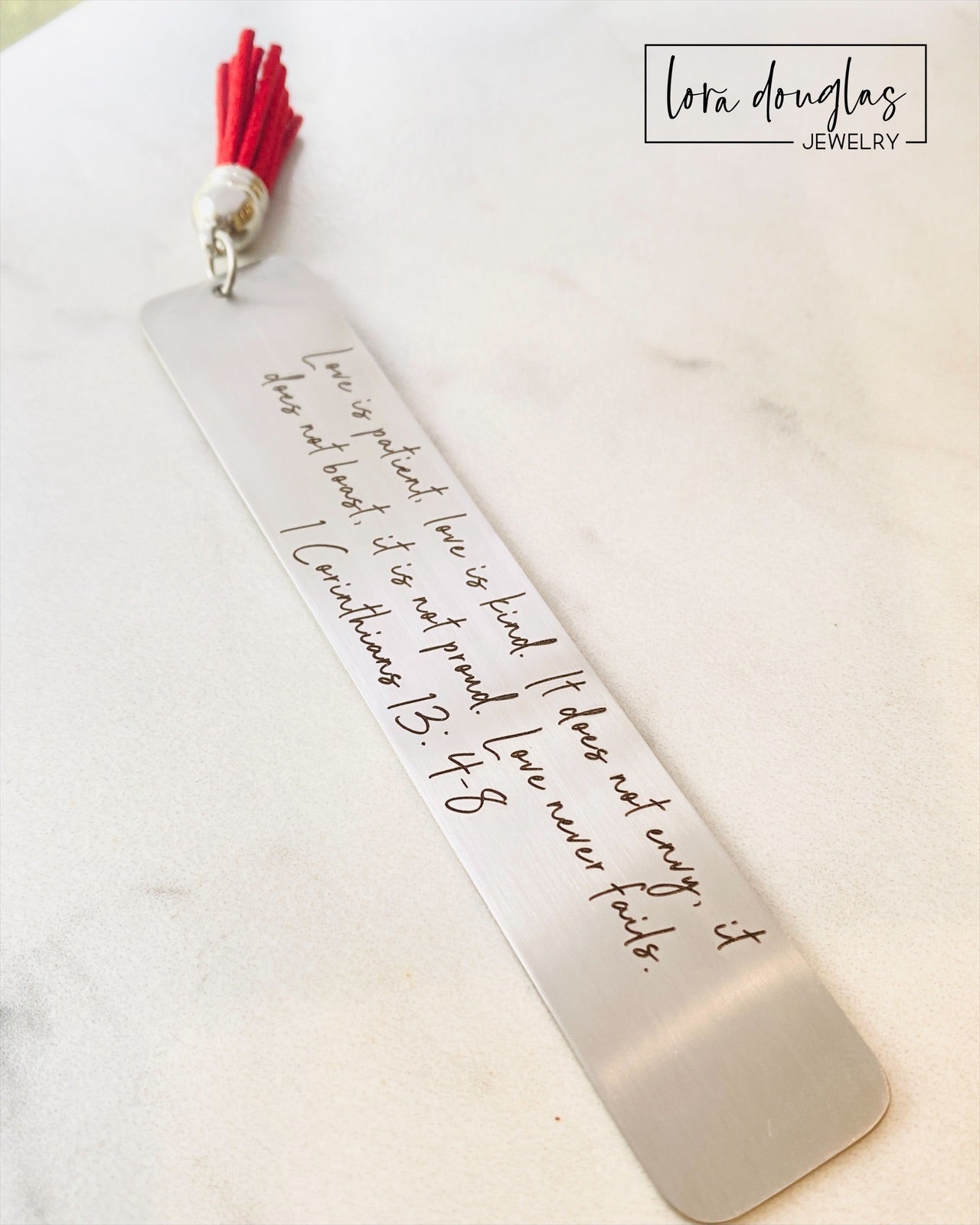 A stainless steel bookmark with the Bible verse 1 Corinthians 13:4-8 engraved on it, attached to a red tassel.