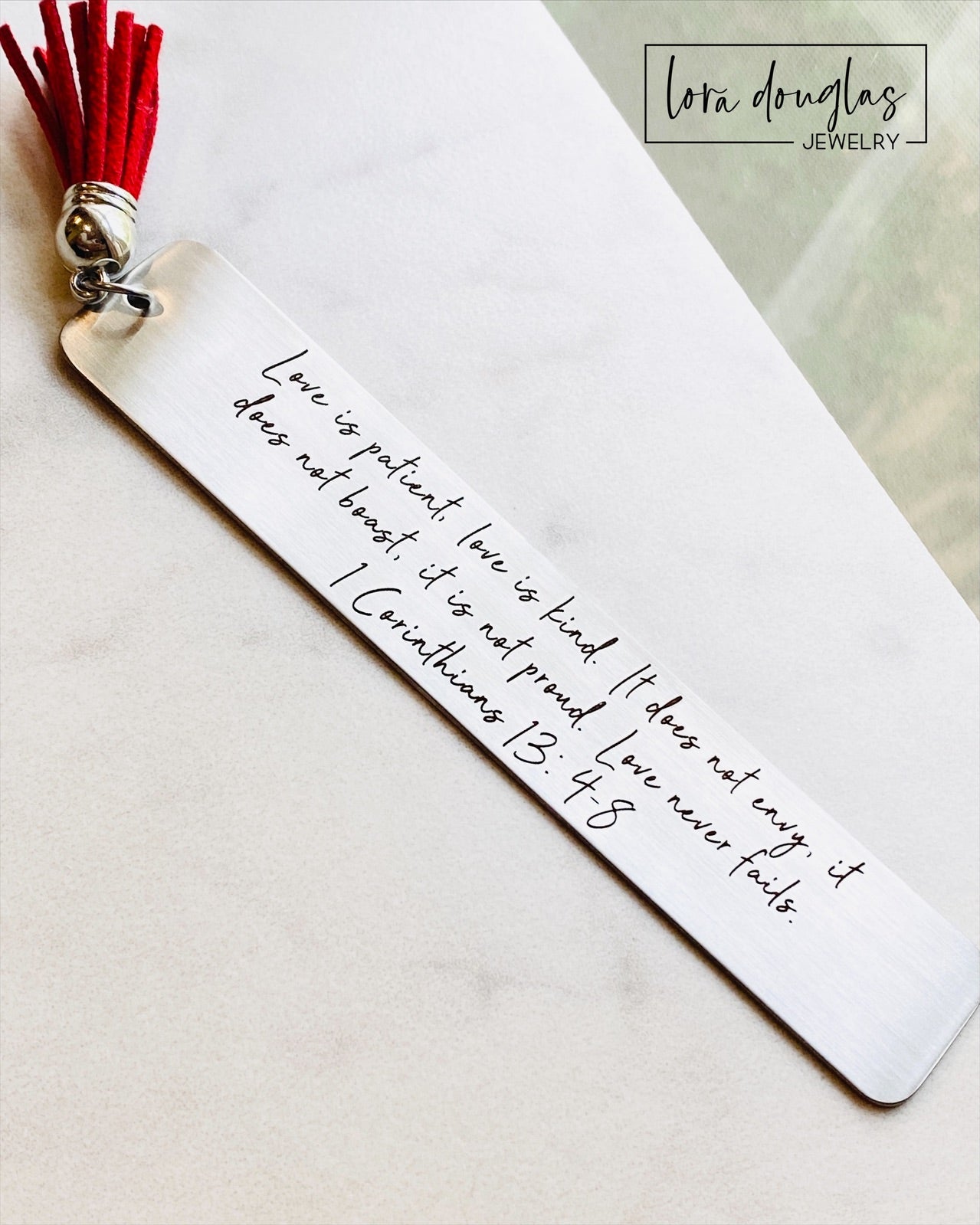 A stainless steel bookmark with the Bible verse 1 Corinthians 13:4-8 engraved on it, attached to a red tassel.