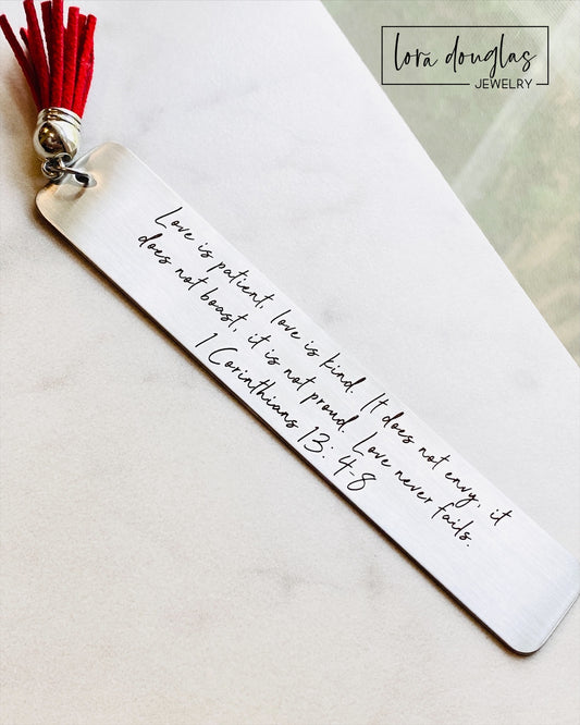 A stainless steel bookmark with the Bible verse 1 Corinthians 13:4-8 engraved on it, attached to a red tassel.