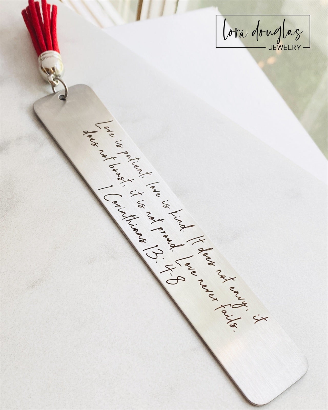 A stainless steel bookmark with the Bible verse 1 Corinthians 13:4-8 engraved on it, attached to a red tassel.