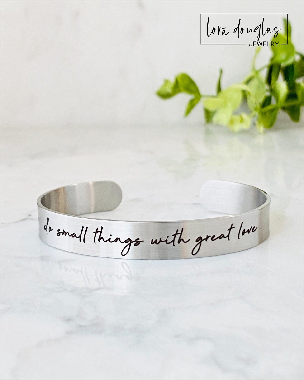 Two stainless steel cuff bracelets, one in silver and one in gold color, each with an engraved message 'Do small things with great love'.