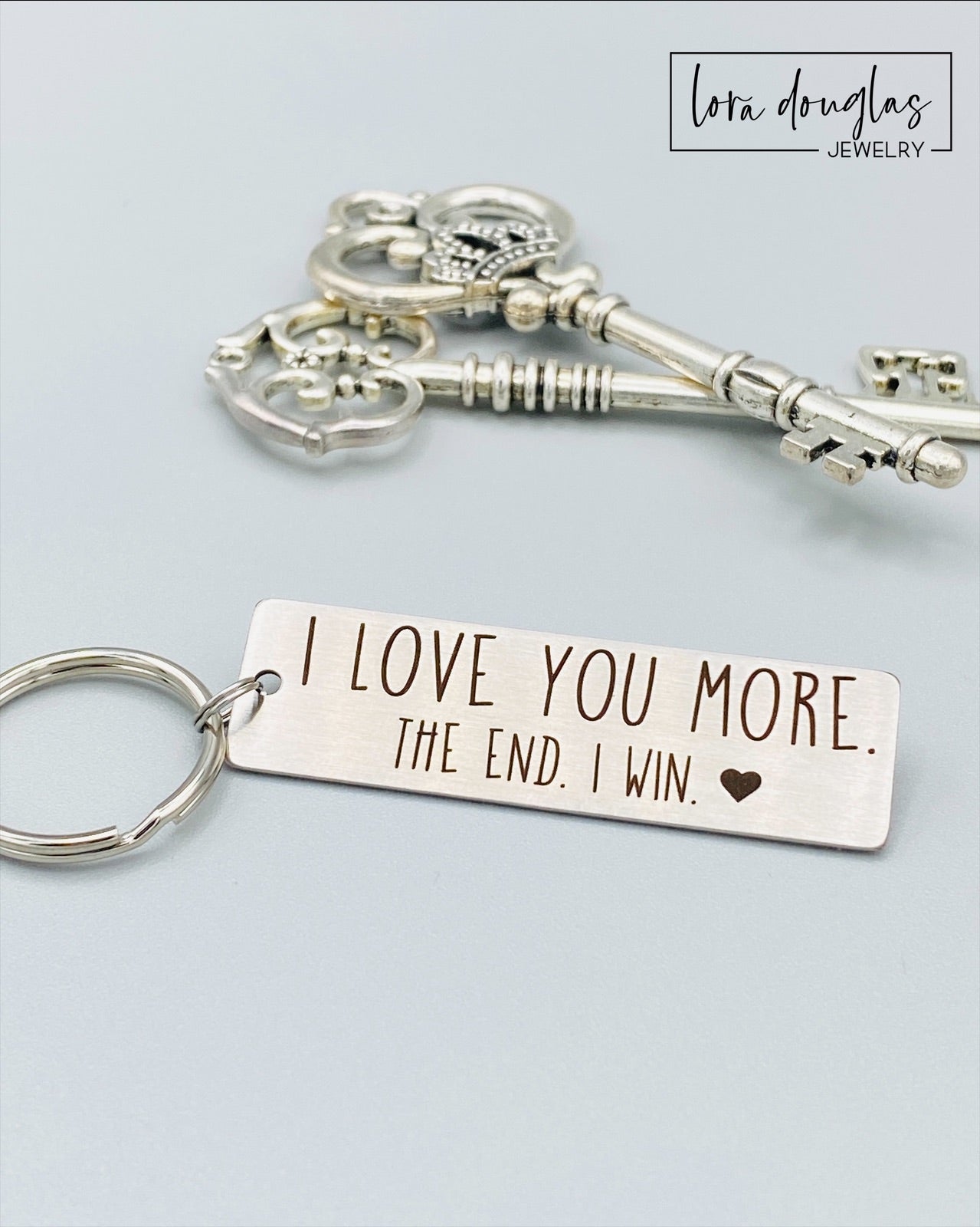 A stainless steel metal keychain with the engraved text 'I Love You More. The End. I Win.' 