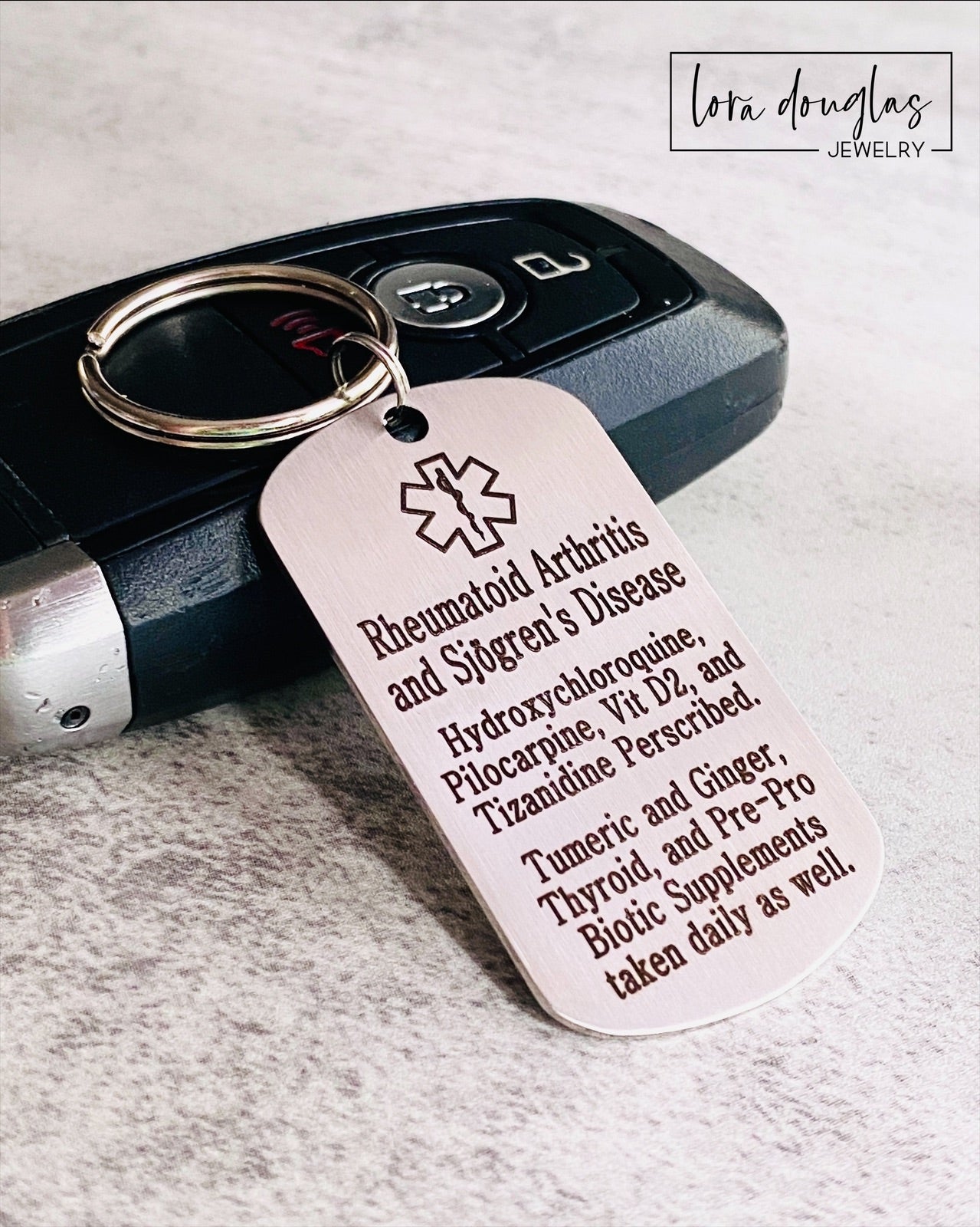 A stainless steel medical alert keychain displayed on a key. The tag has sample text for customization and indicates that additional text can be added on the back.