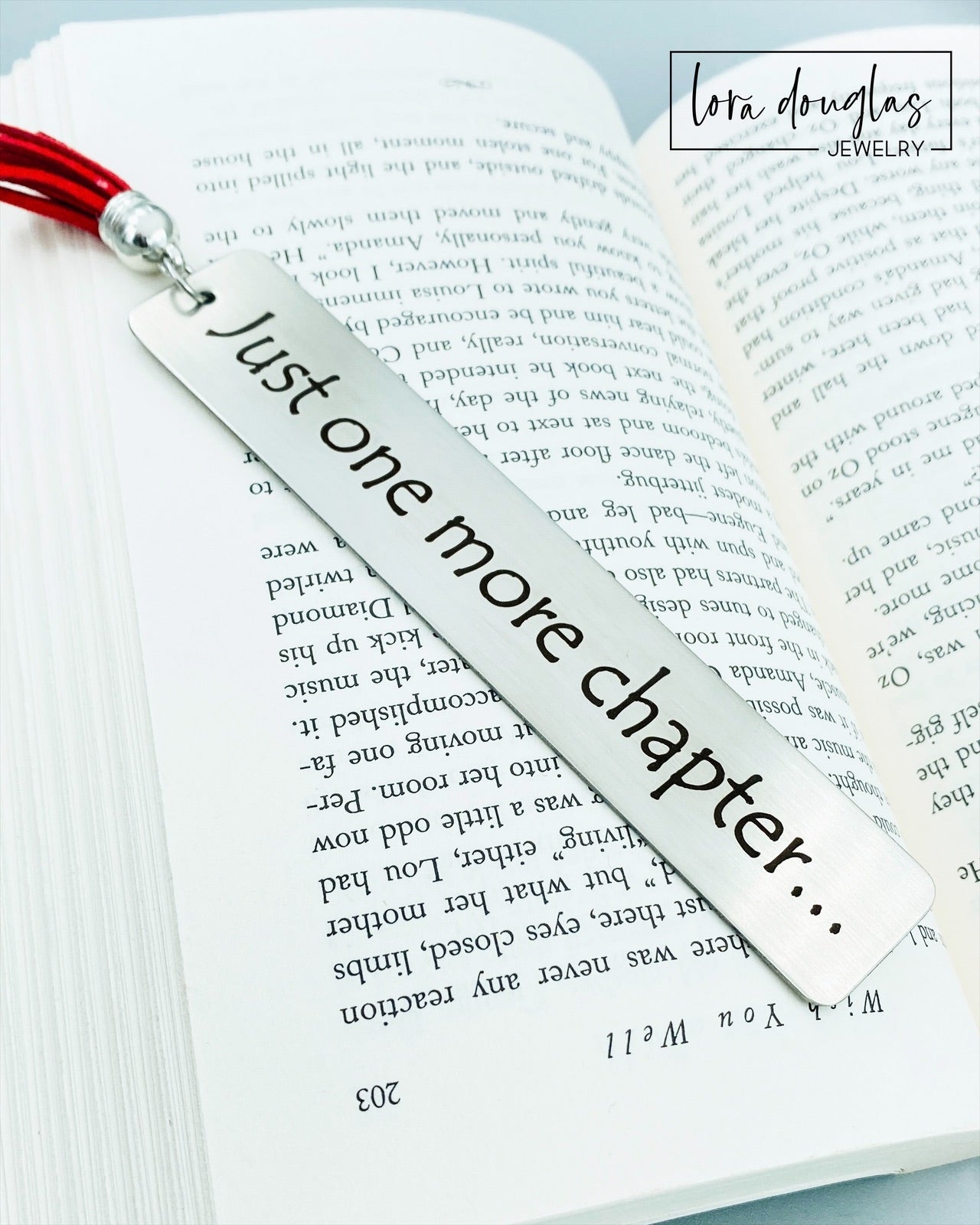 A stainless steel metal bookmark with the engraved text 'Just One More Chapter' and a tassel, placed on an open book.