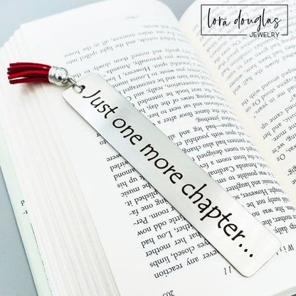 A stainless steel metal bookmark with the engraved text 'Just One More Chapter' and a tassel, placed on an open book.